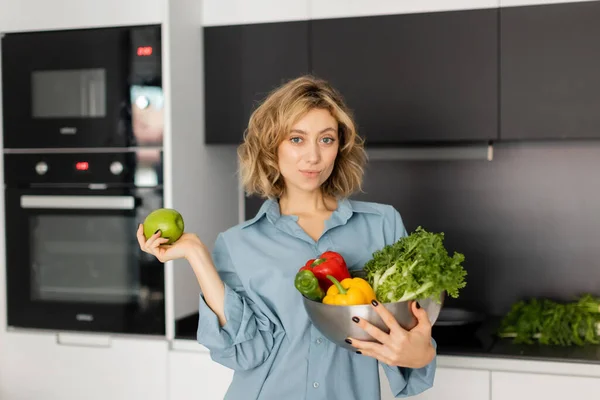 Young woman with wavy hair holding bowl with fresh vegetables and apple in kitchen — Fotografia de Stock