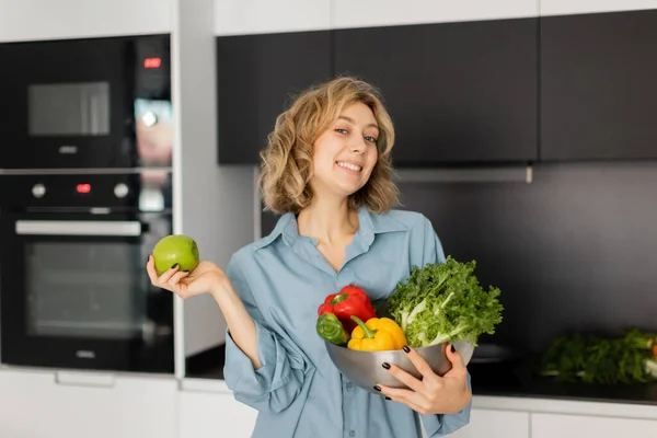 Heureuse jeune femme aux cheveux ondulés tenant bol avec des légumes frais et pomme dans la cuisine — Photo de stock