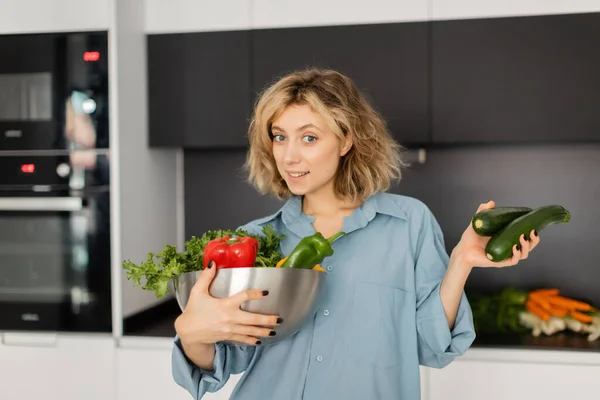 Heureuse jeune femme aux cheveux ondulés tenant bol avec des légumes frais dans la cuisine — Photo de stock