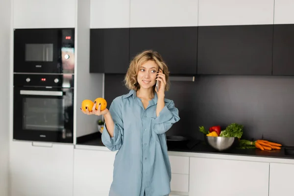 Young woman with wavy hair holding fresh oranges and talking on smartphone in kitchen — Foto stock