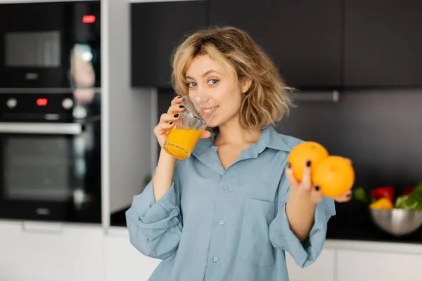 Jovem feliz com cabelo ondulado segurando laranjas frescas e beber suco na cozinha — Fotografia de Stock