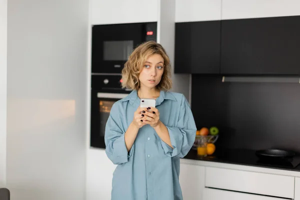 Jovem mulher pensativa com cabelo ondulado segurando smartphone na cozinha — Fotografia de Stock