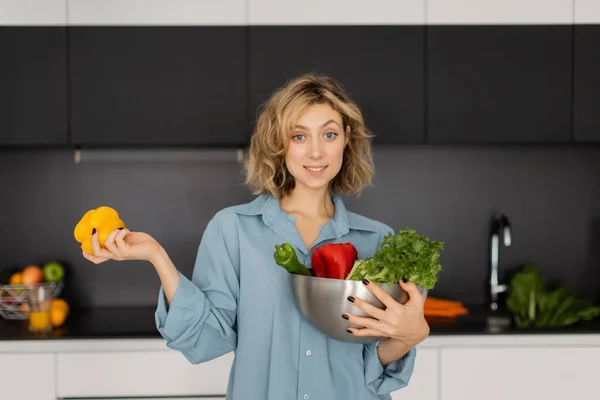Joyeuse jeune femme aux cheveux ondulés tenant bol avec des légumes biologiques dans la cuisine — Photo de stock