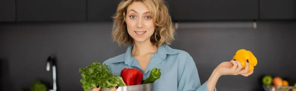 Joyeuse jeune femme aux cheveux ondulés tenant bol avec des légumes biologiques dans la cuisine, bannière — Photo de stock