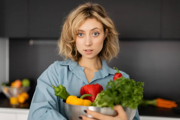 Jovem loira com cabelo ondulado segurando tigela com legumes orgânicos na cozinha — Fotografia de Stock