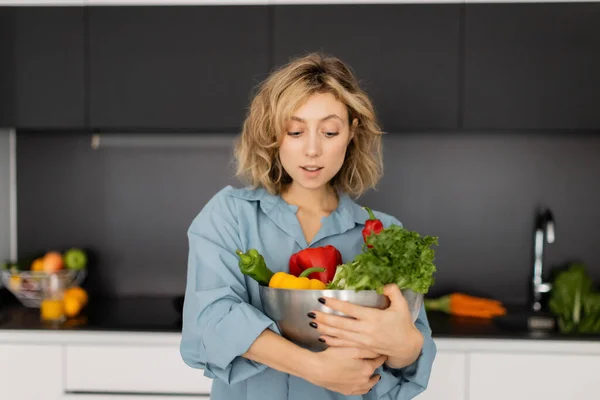 Jolie jeune femme aux cheveux ondulés tenant bol avec des légumes biologiques dans la cuisine — Photo de stock