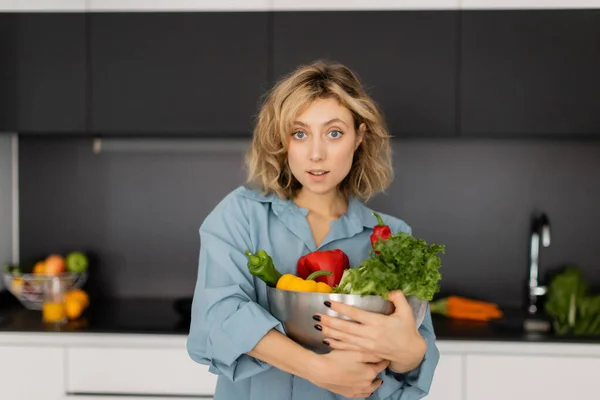 Mulher surpreso e loira com cabelo ondulado segurando tigela com legumes orgânicos na cozinha — Fotografia de Stock