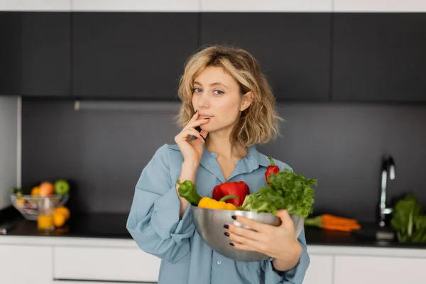 Blonde jeune femme aux cheveux ondulés tenant bol avec des légumes biologiques et regardant la caméra — Photo de stock