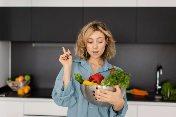Bionda giovane donna con capelli ondulati in possesso di ciotola con verdure biologiche e mostrando segno idea — Foto stock