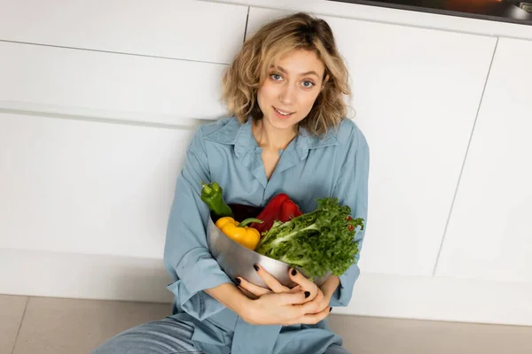 Vue grand angle de jeune femme joyeuse avec des cheveux ondulés tenant bol avec des légumes biologiques dans la cuisine — Photo de stock