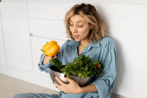 Jovem loira espantada com cabelo ondulado segurando tigela com legumes saborosos na cozinha — Fotografia de Stock