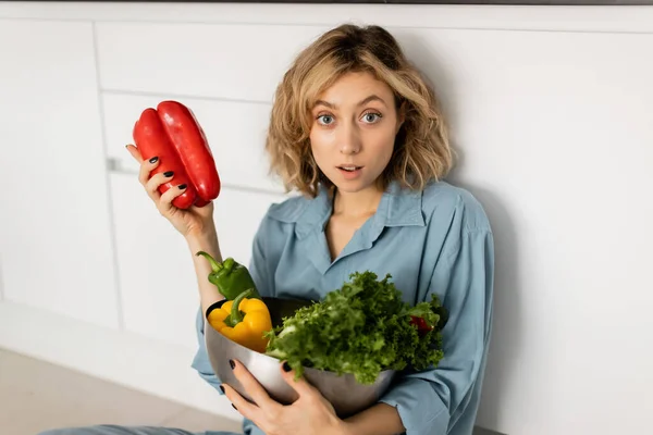Choqué jeune femme aux cheveux ondulés tenant bol avec des légumes biologiques dans la cuisine — Photo de stock