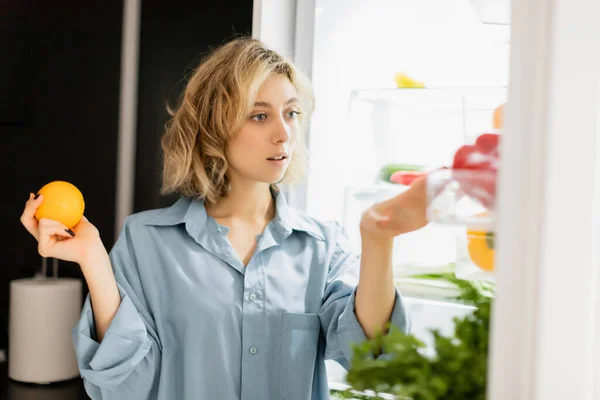 Thoughtful young woman holding orange and looking at refrigerator in kitchen — Photo de stock