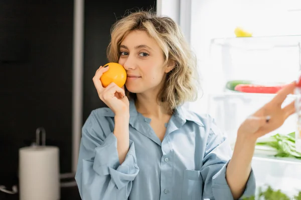 Jovem feliz cheirando laranja perto da geladeira na cozinha — Fotografia de Stock