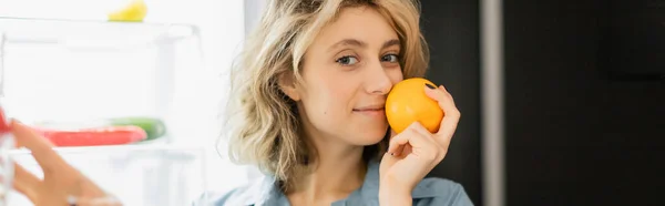 Happy young woman smelling orange near refrigerator in kitchen, banner — Stockfoto