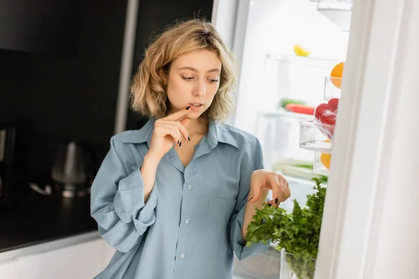 Mujer joven pensativa mirando verdor en refrigerador abierto - foto de stock
