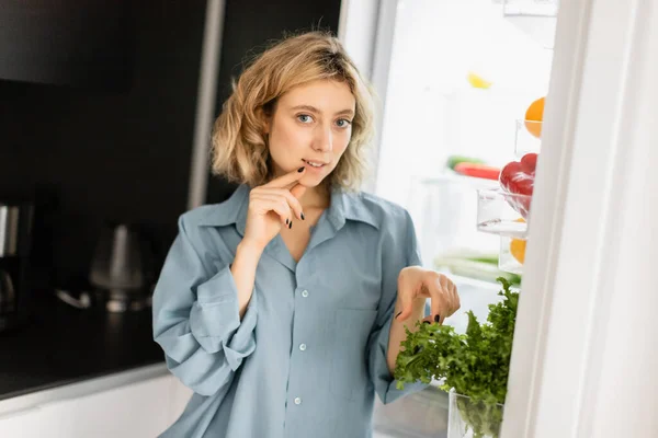 Pensive young woman looking at camera near open refrigerator in kitchen — Stock Photo