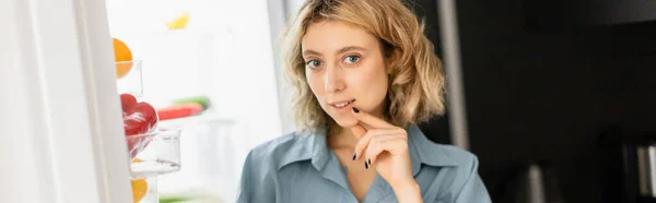 Giovane donna pensierosa che guarda la macchina fotografica vicino al frigorifero aperto in cucina, banner — Foto stock