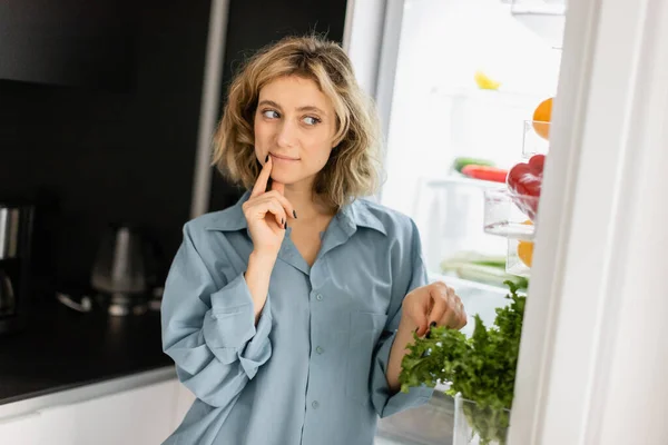 Pensive young woman in blue shirt looking into open refrigerator in kitchen — Stock Photo