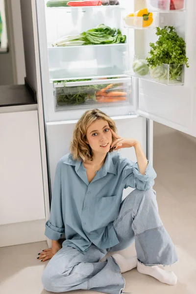 Heureuse jeune femme assise près d'un réfrigérateur ouvert avec des produits frais dans la cuisine — Photo de stock