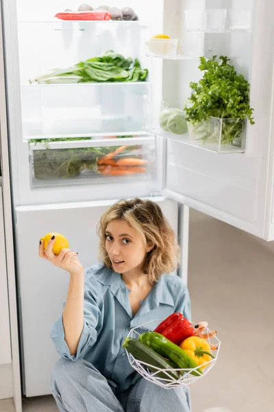 Jovem mulher sentada perto da geladeira aberta e tigela segurando com legumes frescos na cozinha — Fotografia de Stock