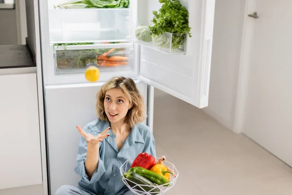 Young woman sitting near opened refrigerator with fresh vegetables while throwing lemon in air - foto de stock