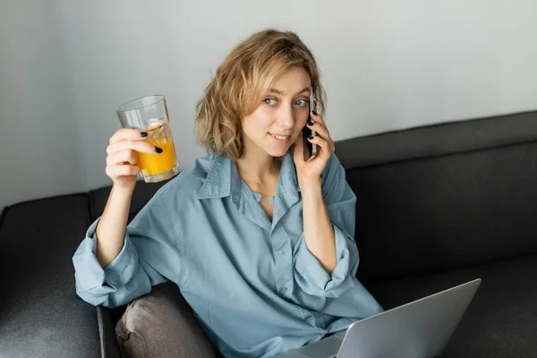 Cheerful freelancer with wavy hair holding glass of orange juice while talking on smartphone near laptop - foto de stock