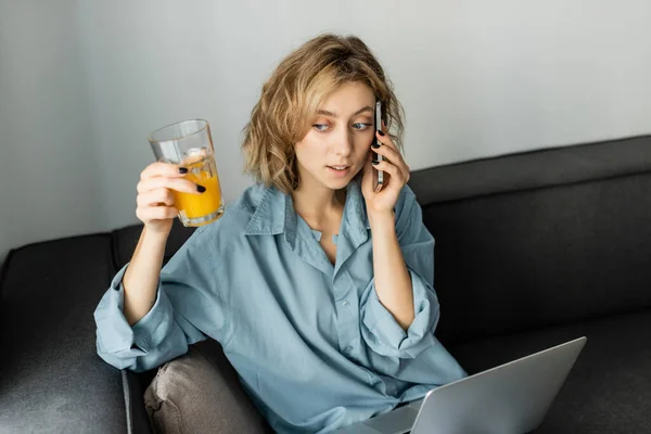 Vista de ángulo alto del joven freelancer con cabello ondulado sosteniendo un vaso de jugo de naranja mientras habla en un teléfono inteligente cerca de la computadora portátil - foto de stock