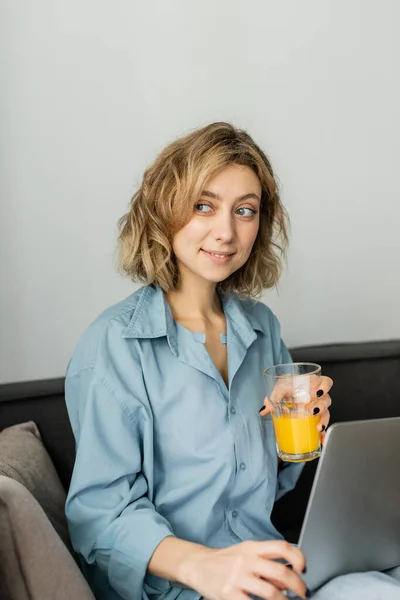Feliz freelancer con cabello ondulado sosteniendo vaso de jugo de naranja cerca de la computadora portátil - foto de stock