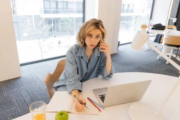 High angle view of young woman talking on smartphone near laptop on desk — Stock Photo