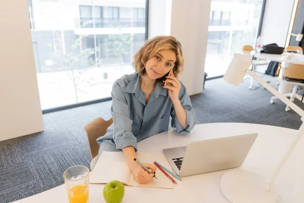 Веселая молодая женщина с волнистыми волосами разговаривает на смартфоне рядом с ноутбуком на столе — стоковое фото