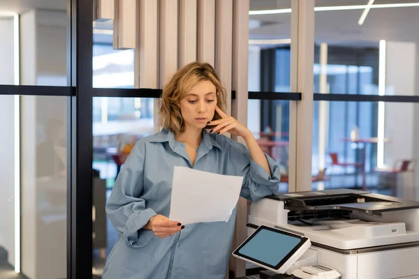 Young blonde woman with wavy hair holding blank paper while standing near printer in office — Photo de stock