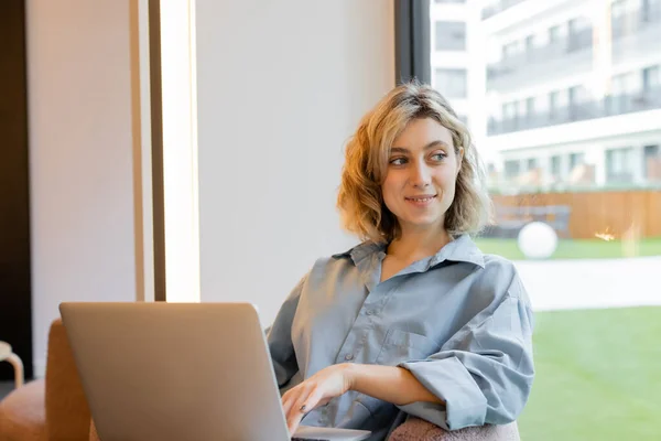 Mujer rubia feliz con el pelo ondulado mirando a la ventana y sentado con el ordenador portátil - foto de stock