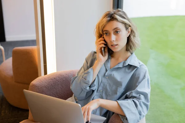 Mujer rubia joven con el pelo ondulado hablando en el teléfono inteligente mientras usa el ordenador portátil en la cafetería - foto de stock