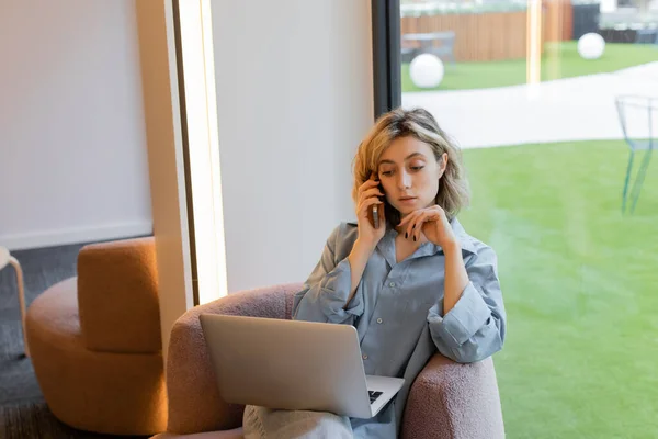 Blonde freelancer with wavy hair talking on smartphone while using laptop in cafe — Stock Photo