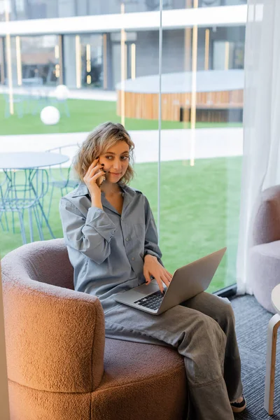 Freelancer alegre com cabelo ondulado falando no smartphone enquanto usa laptop no lobby — Fotografia de Stock