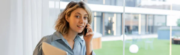 Весела молода жінка з хвилястим волоссям розмовляє на смартфоні і тримає ноутбук біля вікон, банер — стокове фото
