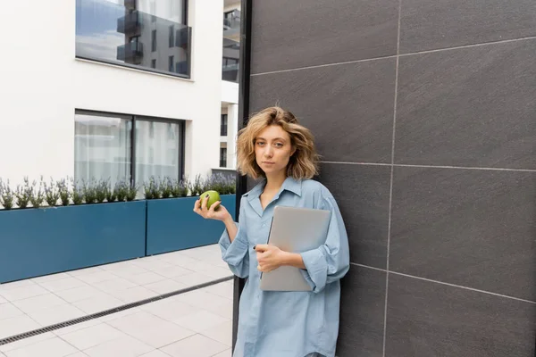 Mujer joven con cabello ondulado sosteniendo manzana verde y portátil cerca del edificio del hotel en Barcelona - foto de stock