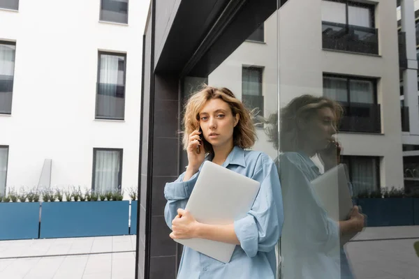 Mujer rubia con el pelo ondulado hablando en el teléfono inteligente y la celebración de la computadora portátil cerca del edificio del hotel en Barcelona - foto de stock
