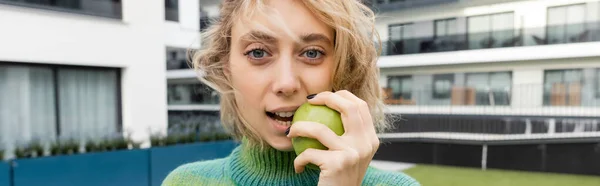 Mujer rubia en suéter comiendo manzana verde cerca del edificio del hotel, pancarta - foto de stock