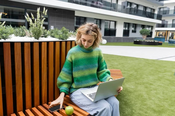 Donna bionda in maglione verde seduta su una panchina di legno con computer portatile mentre prende smartphone vicino all'hotel di Barcellona — Foto stock