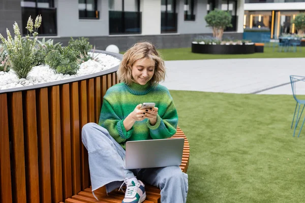 Happy young woman in green sweater sitting on wooden bench with laptop while using smartphone — Foto stock