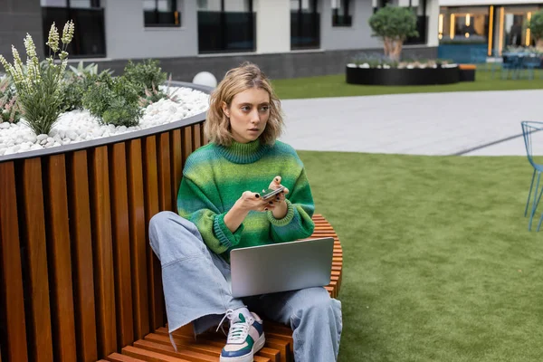 Joven freelancer en jersey verde sentado en un banco moderno con portátil mientras usa el teléfono móvil - foto de stock