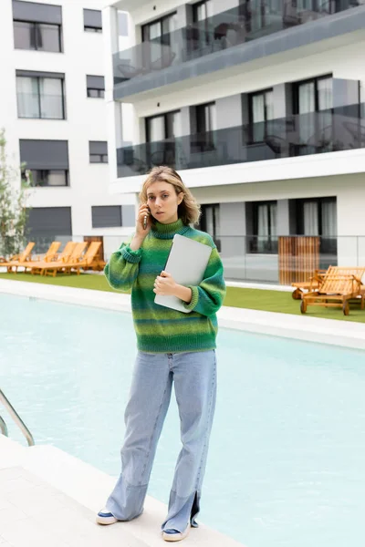 Блондинка у светрі стоїть з ноутбуком, розмовляючи на смартфоні біля відкритого басейну в Барселоні. — стокове фото