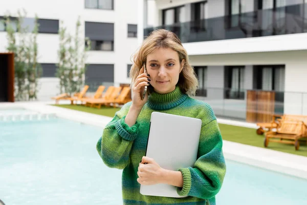 Felice donna in maglione in piedi con computer portatile mentre parla su smartphone vicino alla piscina all'aperto — Foto stock