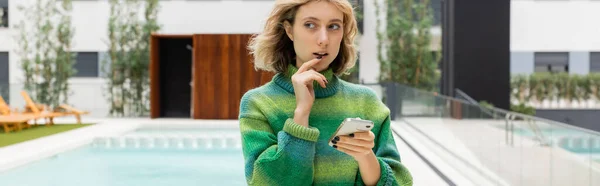 Pensive young woman in sweater holding smartphone near outdoor swimming pool of hotel in Barcelona, banner - foto de stock