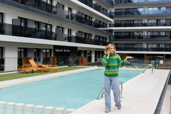 Full length of displeased woman in sweater talking on smartphone near outdoor swimming pool in Barcelona — Stock Photo