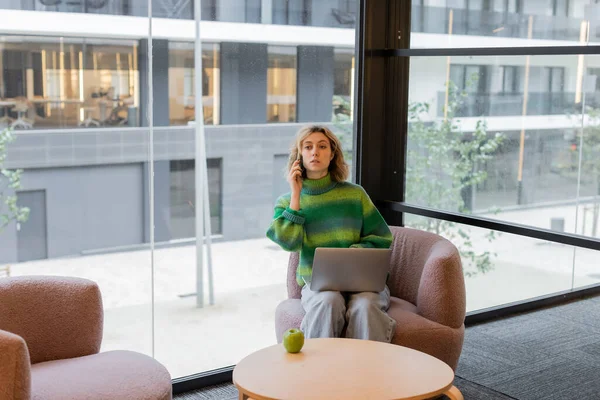 Young woman in sweater talking on smartphone while sitting with laptop in lobby of hotel in Barcelona — Stock Photo