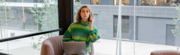 Mujer joven en suéter hablando en el teléfono inteligente mientras se sienta con el ordenador portátil en el vestíbulo del hotel en Barcelona, pancarta - foto de stock