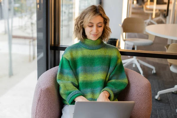 Cheerful young woman in sweater using laptop in lobby of hotel in Barcelona — Stock Photo